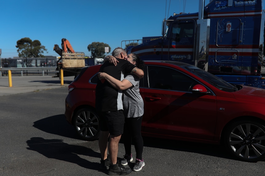 Sean Mulvaney hugs his partner, Liz, goodbye at a petrol station in front of a red car on a sunny day.