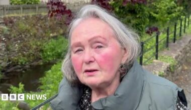 Rhona Barton has grey hair and is looking at the camera. She is wearing a green sleeveless jacket and is standing by a river which is fenced off.