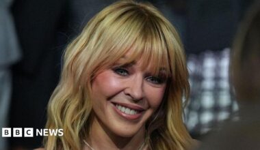 A close-up portrait of singer Kylie Minogue smiling. She has blonde, shoulder-length hair with a full fringe. She is wearing a pearl necklace. The background is blurred with dark tones and a grid pattern.