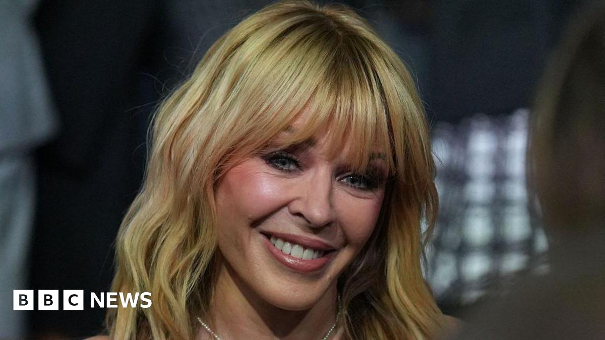 A close-up portrait of singer Kylie Minogue smiling. She has blonde, shoulder-length hair with a full fringe. She is wearing a pearl necklace. The background is blurred with dark tones and a grid pattern.