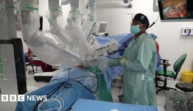 A robot with several arms performing a surgery in an operating theatre. There is a surgeon in a green frock coat who is wearing a face mask. There is a monitor in the background and surgical equipment in the foreground.