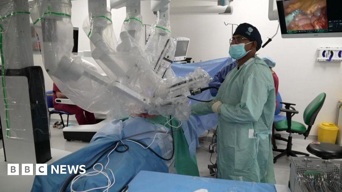 A robot with several arms performing a surgery in an operating theatre. There is a surgeon in a green frock coat who is wearing a face mask. There is a monitor in the background and surgical equipment in the foreground.