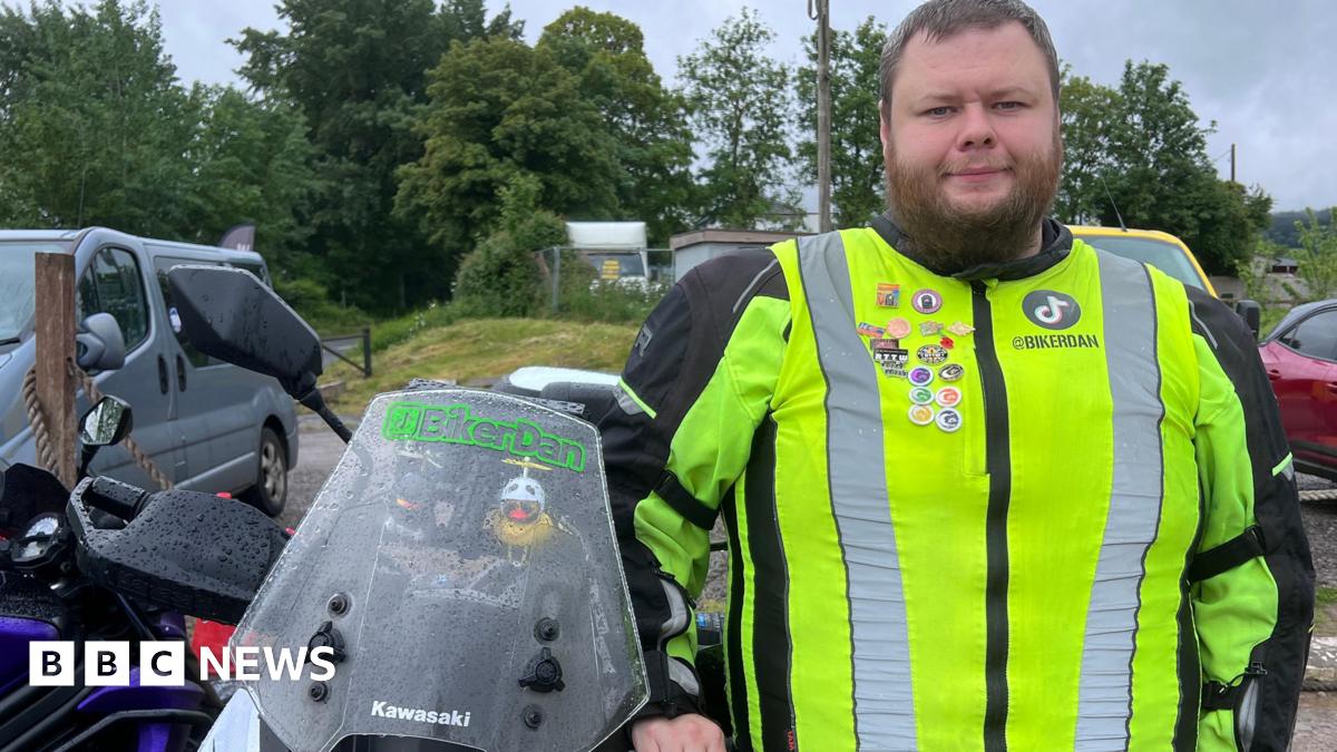 A man with a beard in a high vis jacket stands next to a motorbike in a car park with trees in the background. He has "BikerDan" written on the windshield of his motorbike.