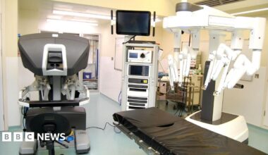 A hospital theatre with a robot with several "arms" above a surgery bed. There are a number of other machines in the room, as well as medicines and cleaning supplies.