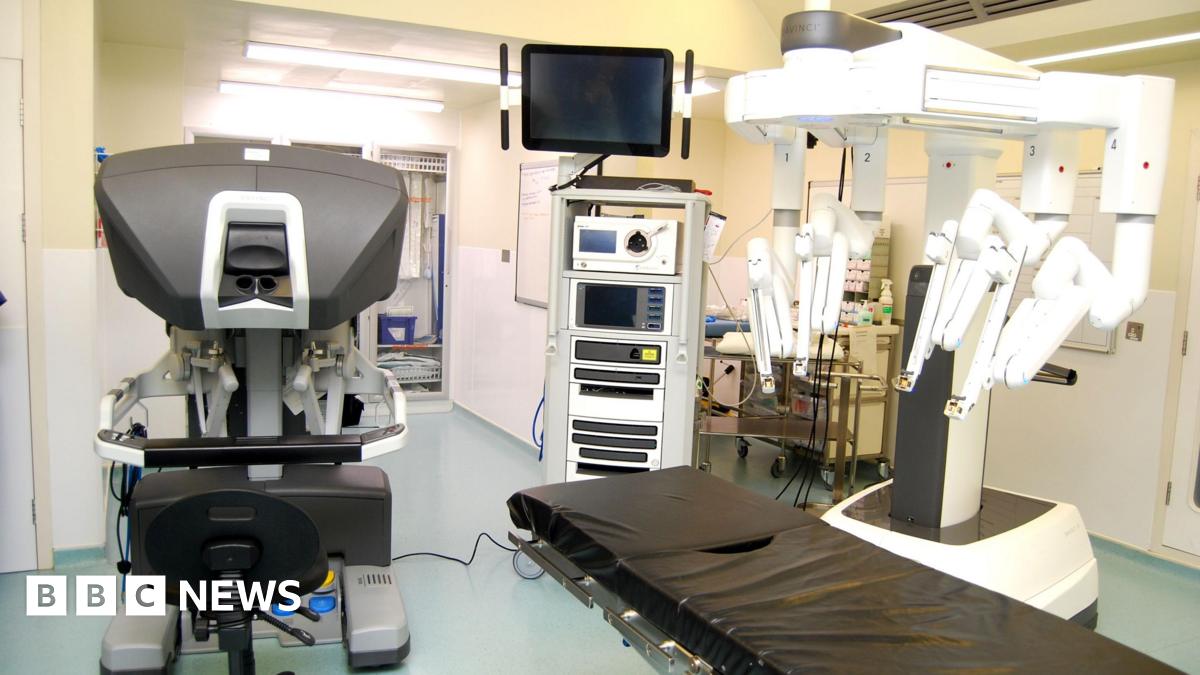 A hospital theatre with a robot with several "arms" above a surgery bed. There are a number of other machines in the room, as well as medicines and cleaning supplies.