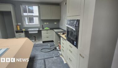 An olive green kitchen with black worktops and large gold handles which is half-renovated and built with protective polystyrene and cardboard around some parts of it. There is a window at the back above the sink.
