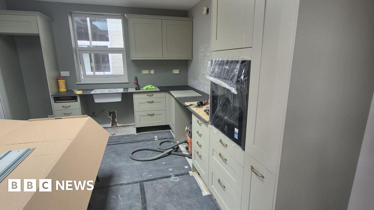 An olive green kitchen with black worktops and large gold handles which is half-renovated and built with protective polystyrene and cardboard around some parts of it. There is a window at the back above the sink.