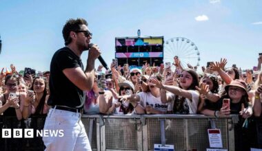 Niall Horan performs at Radio 1's Big Weekend in Sunderland wearing sunglasses, white jeans and black t-shirt. Young female fans behind metal barrier with phones in their hands reach out towards him, arms outstretched.  A Ferris wheel is pictured in the background against a blue sky.