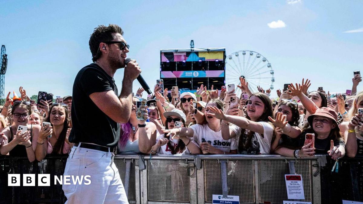 Niall Horan performs at Radio 1's Big Weekend in Sunderland wearing sunglasses, white jeans and black t-shirt. Young female fans behind metal barrier with phones in their hands reach out towards him, arms outstretched.  A Ferris wheel is pictured in the background against a blue sky.