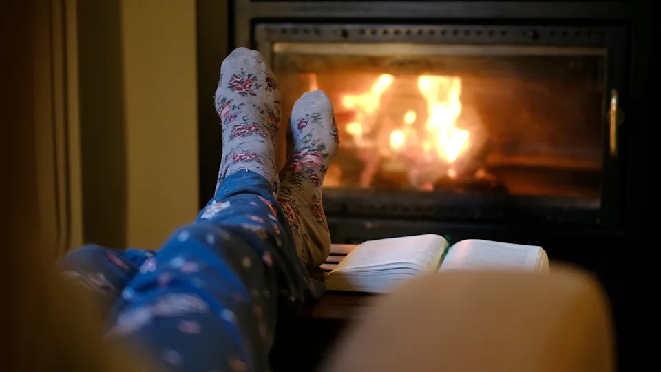 A person wearing patterned socks and blue pajamas relaxes with feet propped up near a cozy fireplace, a book lying open beside them
