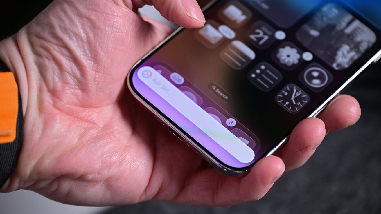 Hand holding a smartphone displaying a dark home screen with widgets and a bright purple Siri search bar at the bottom, with fingers resting near the phone's edge