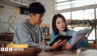 A young couple sit in their kitchen looking at bills. The woman is holding papers and the man is holding a tablet device