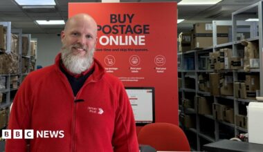 Chris wearing his red Jersey Post fleece jacket standing in front of the counter at Jersey Post headquarters. A sign behind him says "Buy postage online: Save time and skip the queues."