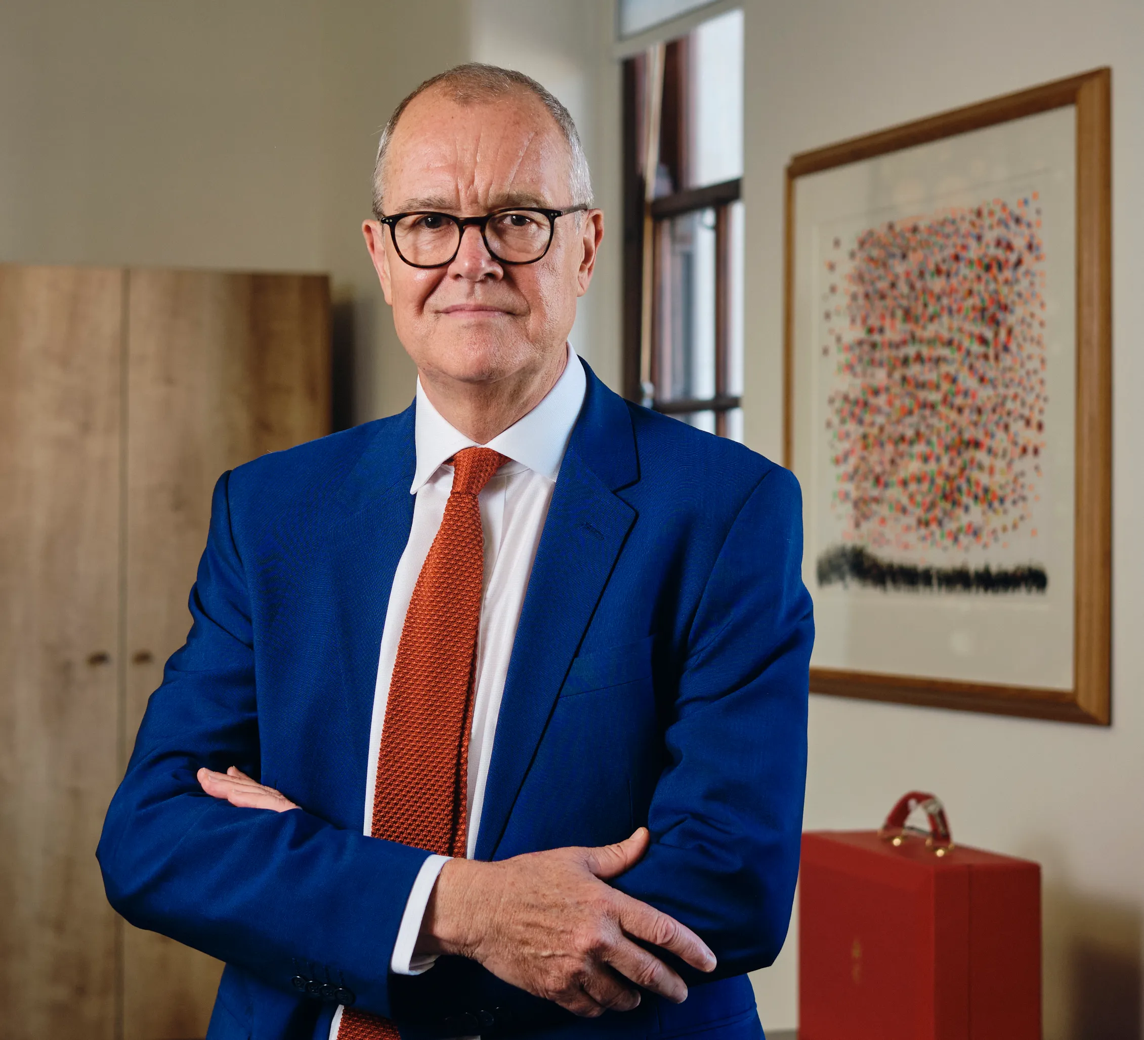 Lord Patrick Vallance, a peer and scientist, standing in a blue suit with an orange tie, arms crossed, looking at the viewer.