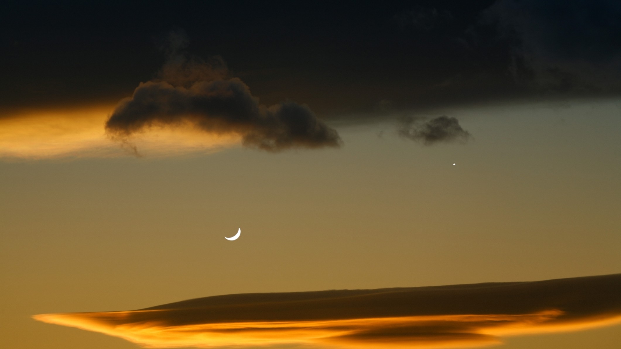 A crescent moon shines to the left of Venus, they are surrounded above and below by lenticular clouds, glowing golden by from setting sun below.