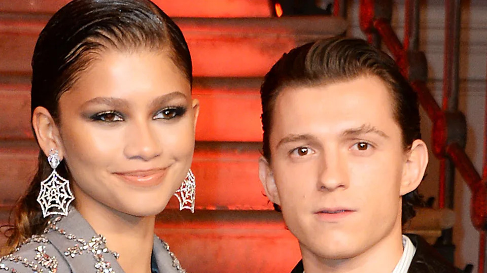 Zendaya and Tom Holland pose together, Zendaya wearing spider web earrings, both with slicked-back hair, in front of red-lit stairs.
