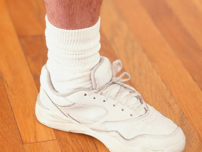Person's leg with a visible scar walking on a wooden floor, wearing white sneakers and socks