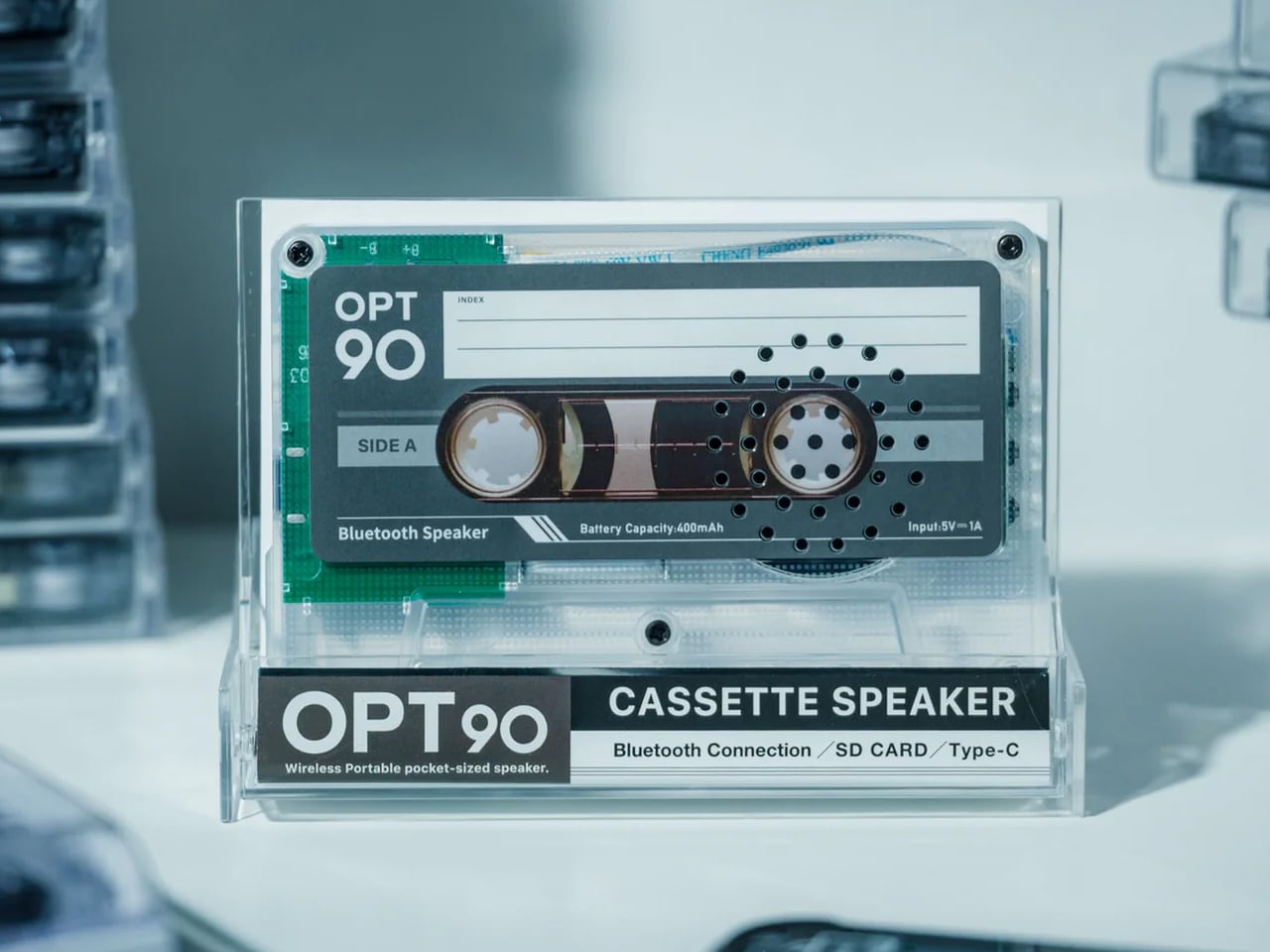 Transparent display of an OPT90 cassette speaker in a clear case, with 'CASSette SPEAKER' and 'Bluetooth Connection' labels visible.