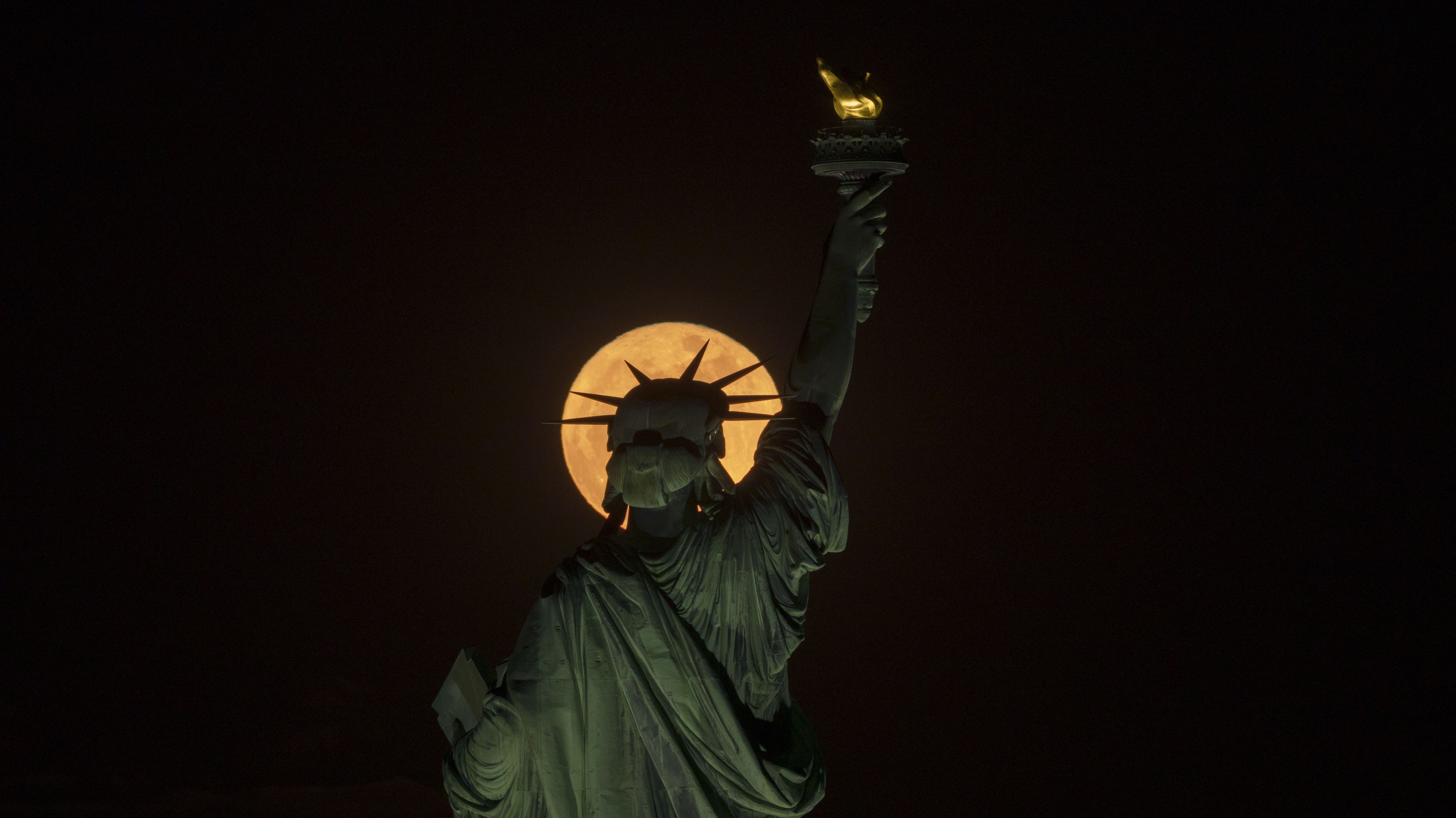 A yellow full moon is pictured rising behind the statue of liberty at night, making it appear as if the lady of liberty is wearing a golden halo.
