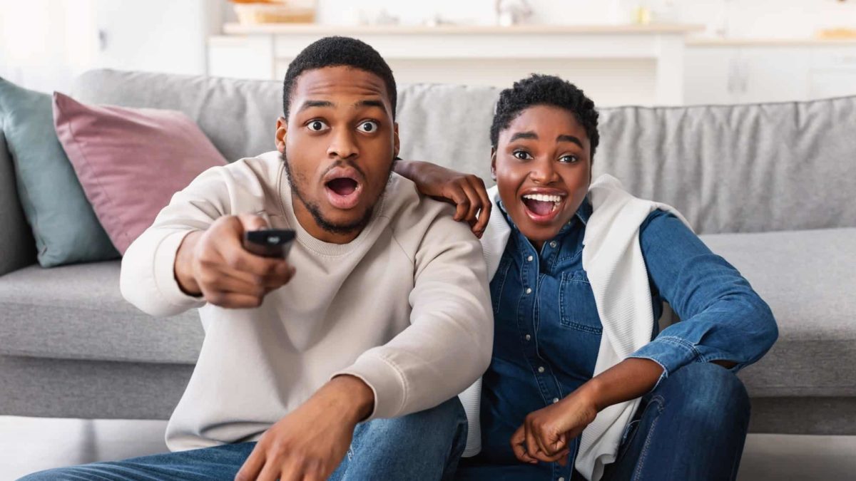 A couple stares at the tv in shock, with the man holding the remote up ready to press a button.