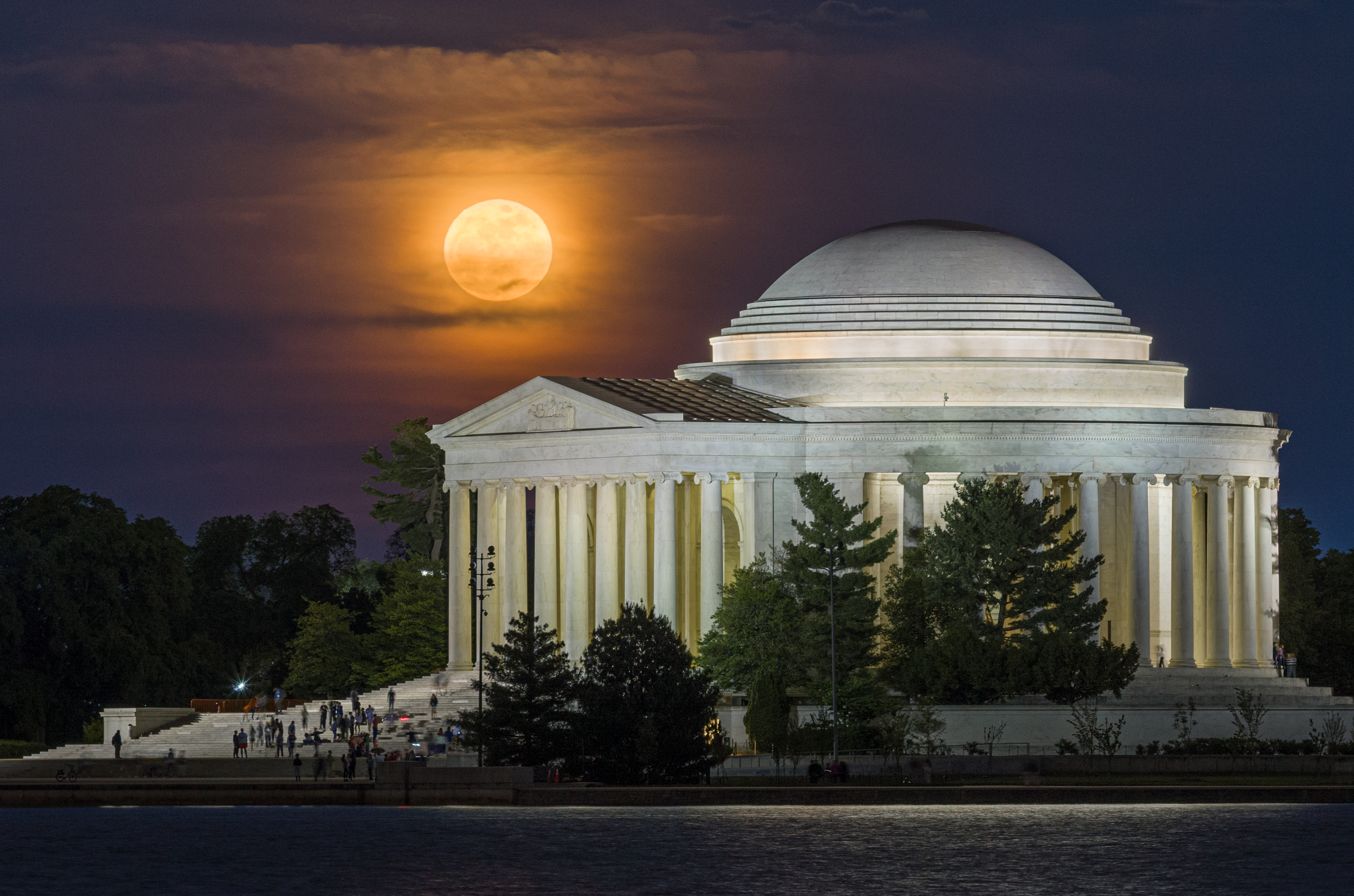 An orange full moon illuminates thin clouds above the domed exterior of the Jefferson Memorial in Washington D.C. at night. Trees are visible in the foreground bordering a lake.
