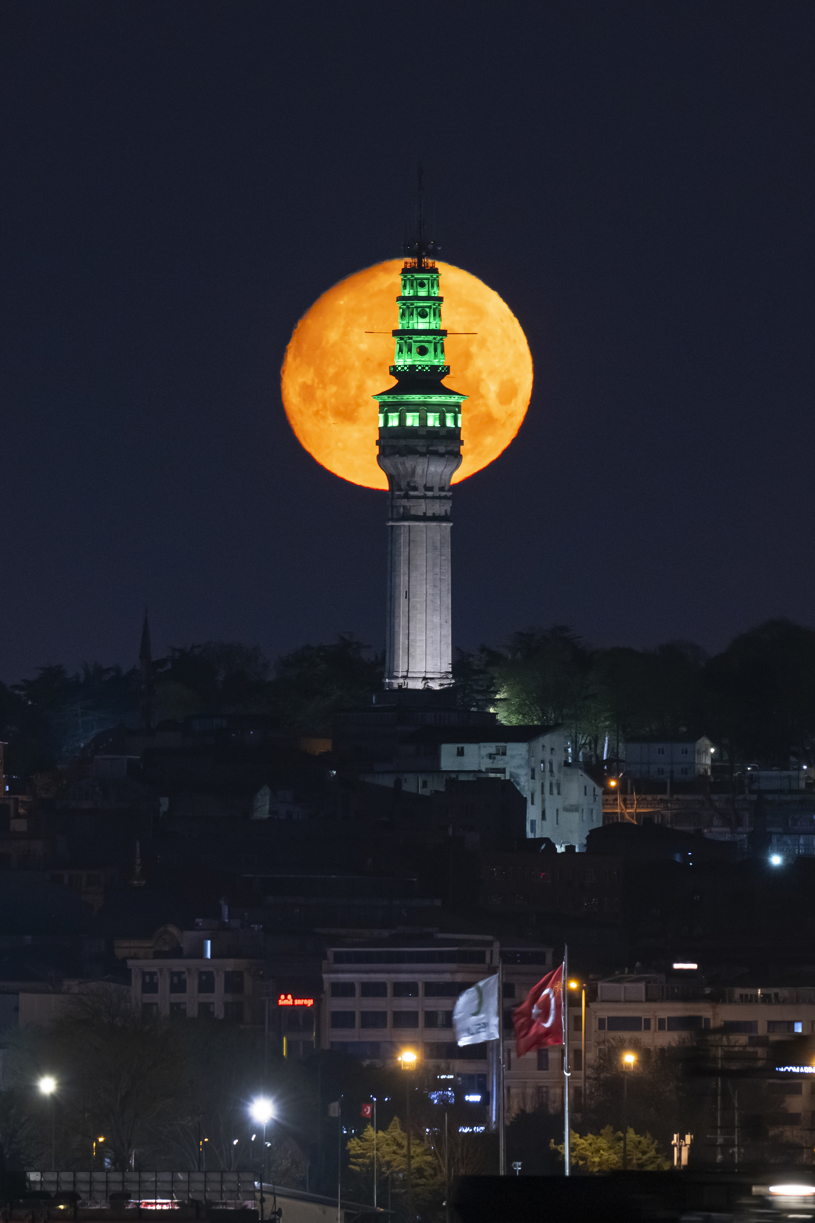 A yellow-orange full moon is pictured rising behind a white tower on a hillside at night. The top section of the tower is lit green.