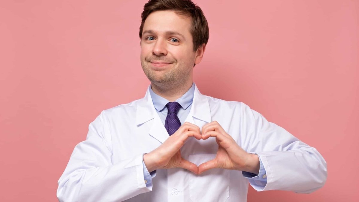 A doctor in a white coat makes a heart shape with his hands and holds it over his chest where his heart is placed.
