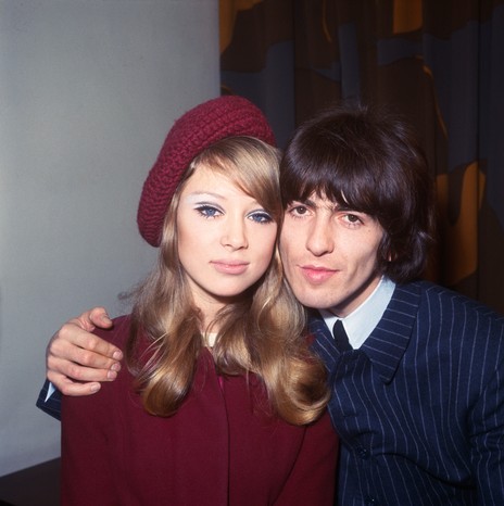 George Harrison and Pattie Boyd in 1966 Colour photo of a young couple (Pattie Boyd and George Harrison) posing closely together indoors. The woman, wearing a red beret and matching coat, has long blonde hair and looks directly at the camera with a neutral expression. The man, in a dark pinstripe suit, leans in with his arm around her shoulders, also facing the camera. A plain wall and dark curtain form the background.
