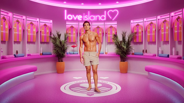 ©ITV A man stood in a pink room wearing tanned swim shorts with one hand in his pocket.