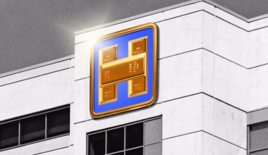 A black and white photo of the side of a hospital building with a large, color sign of an H made of gold bars on a blue background with a gold rim.