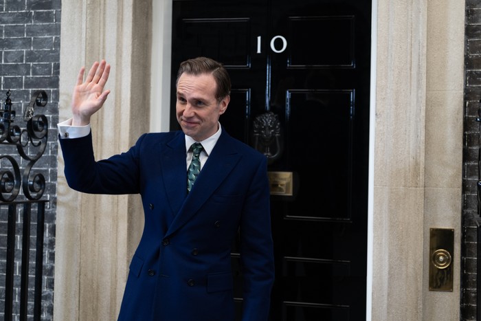 Number 10 First Look Rafe Spall as Prime Minister Harry Douglas in Number 10, wearing a suit, stood outside 10 Downing Street, waving.