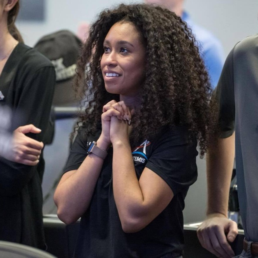 A woman with gorgeous curly hair holds her hands clasped at her chest, and looks to the left with a hopeful, awe-inspired smile.