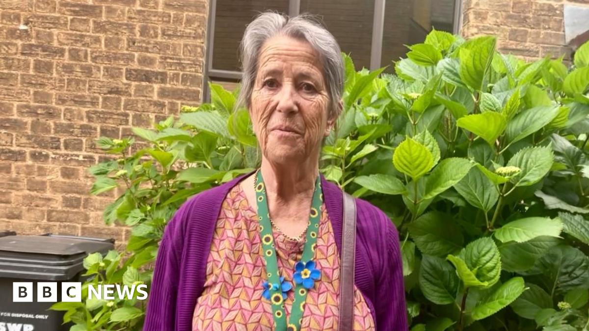 A woman in her 60s with long grey hair is wearing a purple cardigan and orange patterned dress. She is standing in her garden.