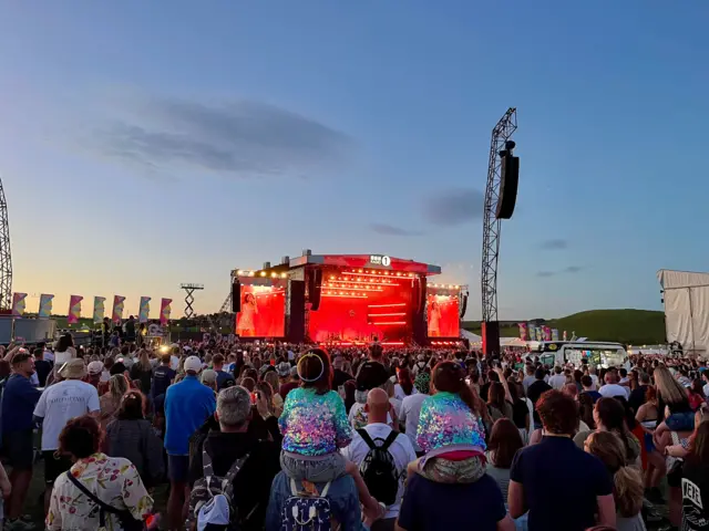 A view from the back of the crowd looking towards the stage which consists of the main stage and two large screens at either side showing Olivia Dean. The stage is bathed in red light. In the foreground are two children on two men's shoulderss