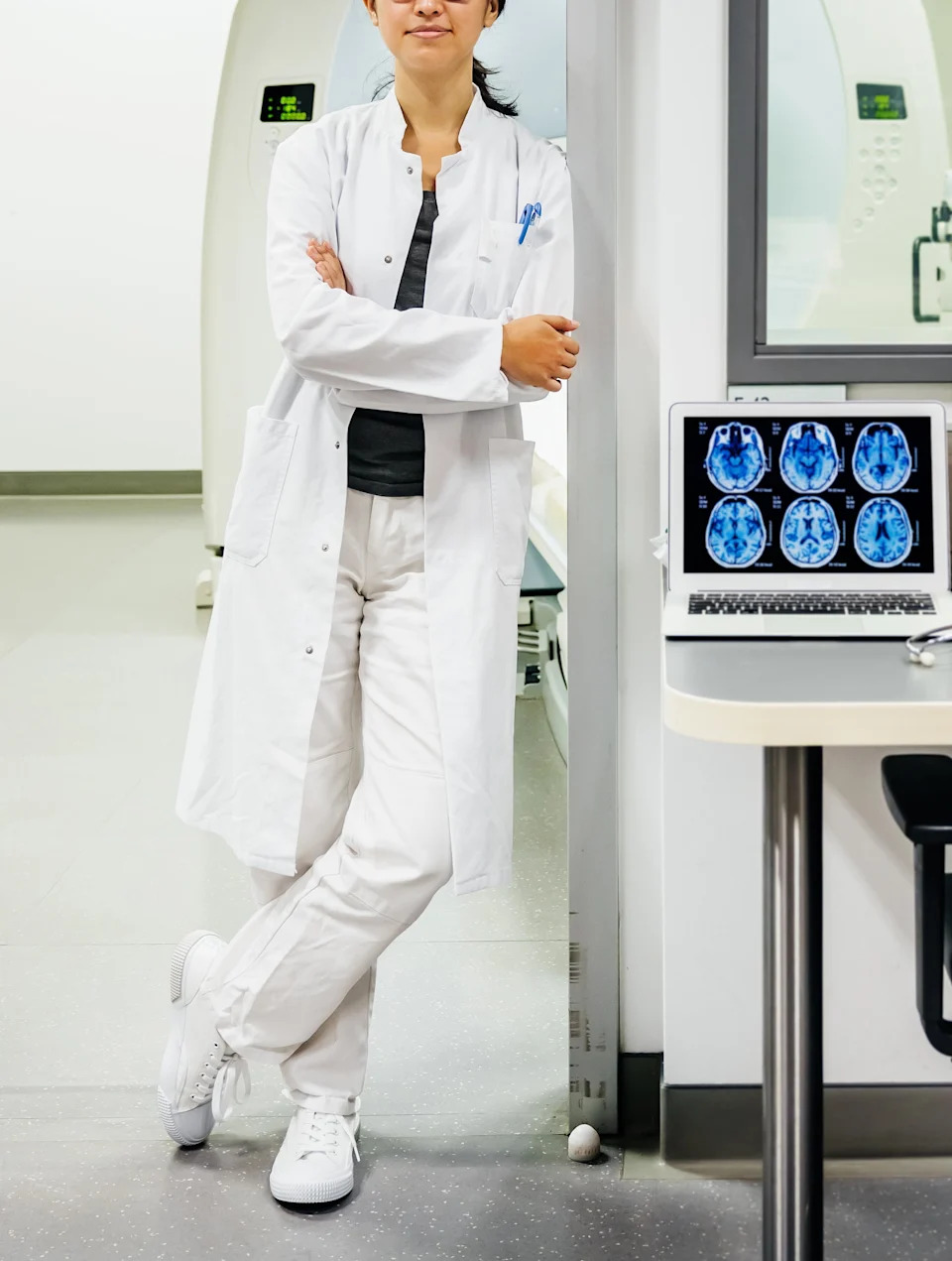 Person in a lab coat stands in a medical research lab, next to brain scan equipment, conveying a professional and scientific environment