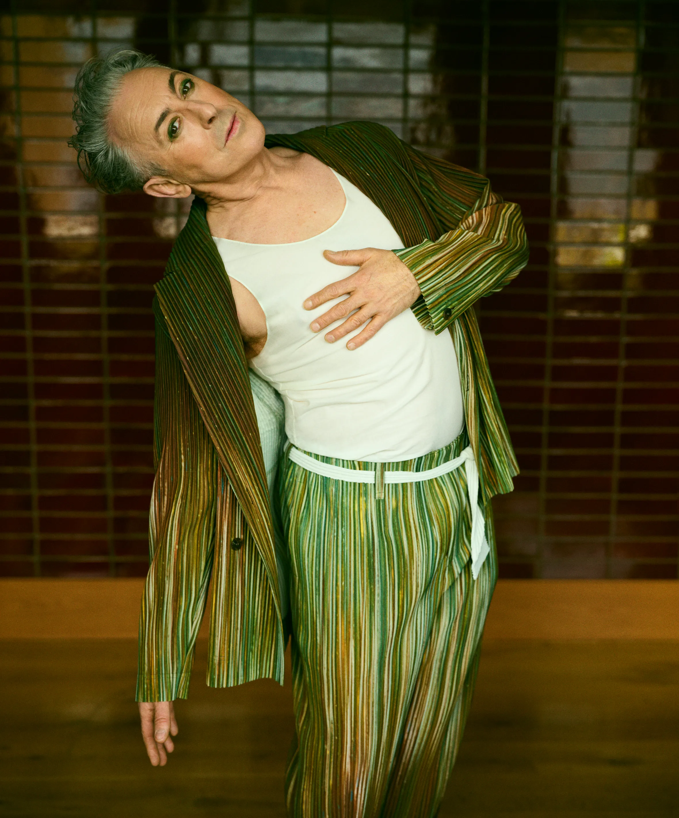 Alan Cumming in a vertical striped green, brown, and white suit, with a white tank top and a matching belt, bending sideways with his head tilted.