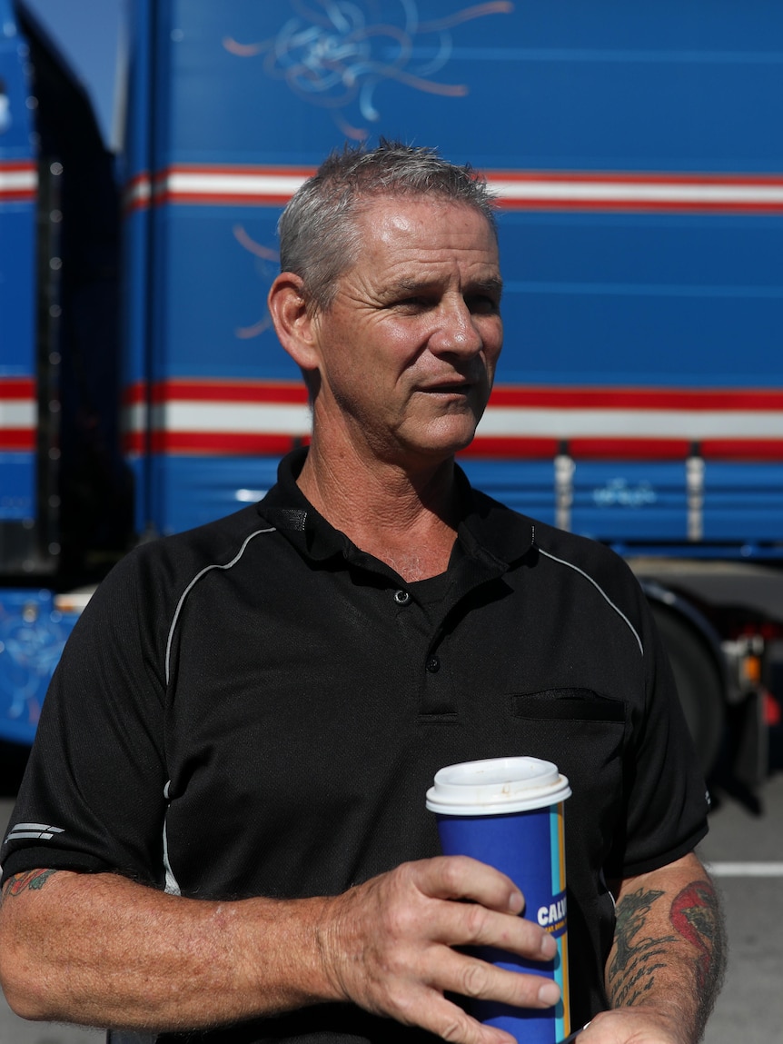 A man in a black polo holds a large coffee cup with a massive blue and red truck seen behind him.