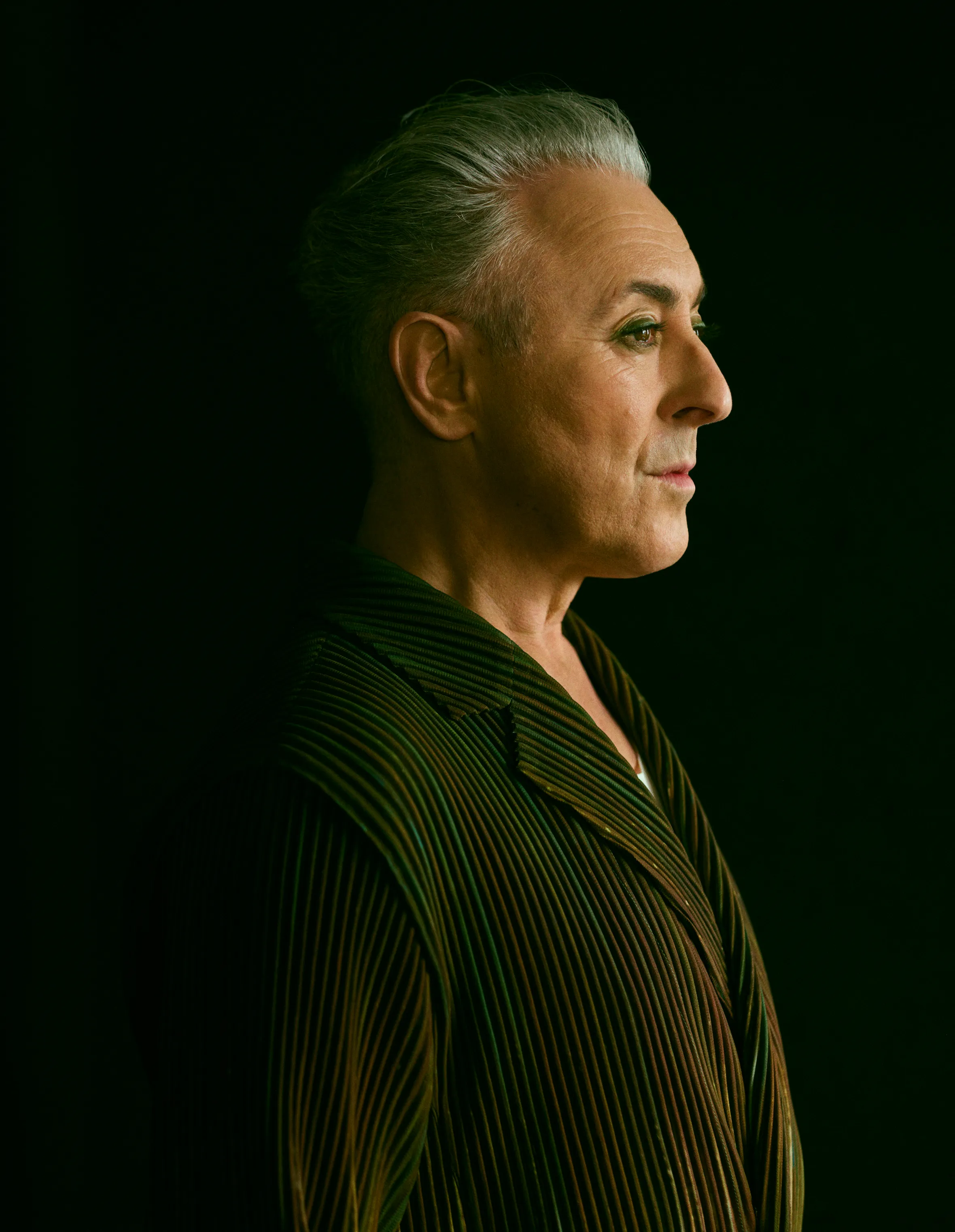 Profile shot of Alan Cumming with gray hair and green eyeshadow, wearing a green striped jacket.