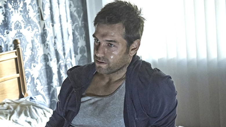 Lucas hood wearing a grey shirt and blue jacket sitting on a bed with sweat on his face in 
