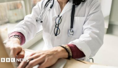 A stock image of a doctor working on a computer - the image does not show her head, she is shown from the shoulders down from an angle behind the computer she is working at. She is typing, wearing a white coat with a stethescope draped around her neck and black-rimmed glasses hanging from the front of the coat. Her hands in the foreground are typing on the keyboard.