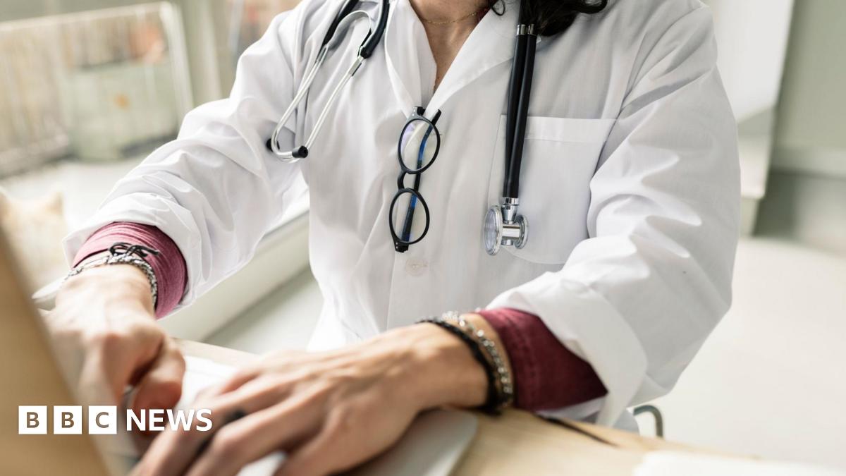 A stock image of a doctor working on a computer - the image does not show her head, she is shown from the shoulders down from an angle behind the computer she is working at. She is typing, wearing a white coat with a stethescope draped around her neck and black-rimmed glasses hanging from the front of the coat. Her hands in the foreground are typing on the keyboard.