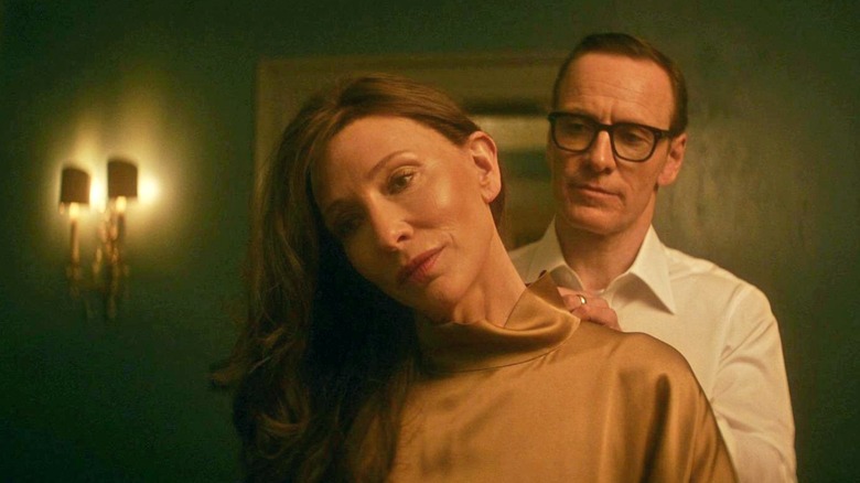 George clasps a necklace around the neck of Kathryn in a dimly-lit hallway in 