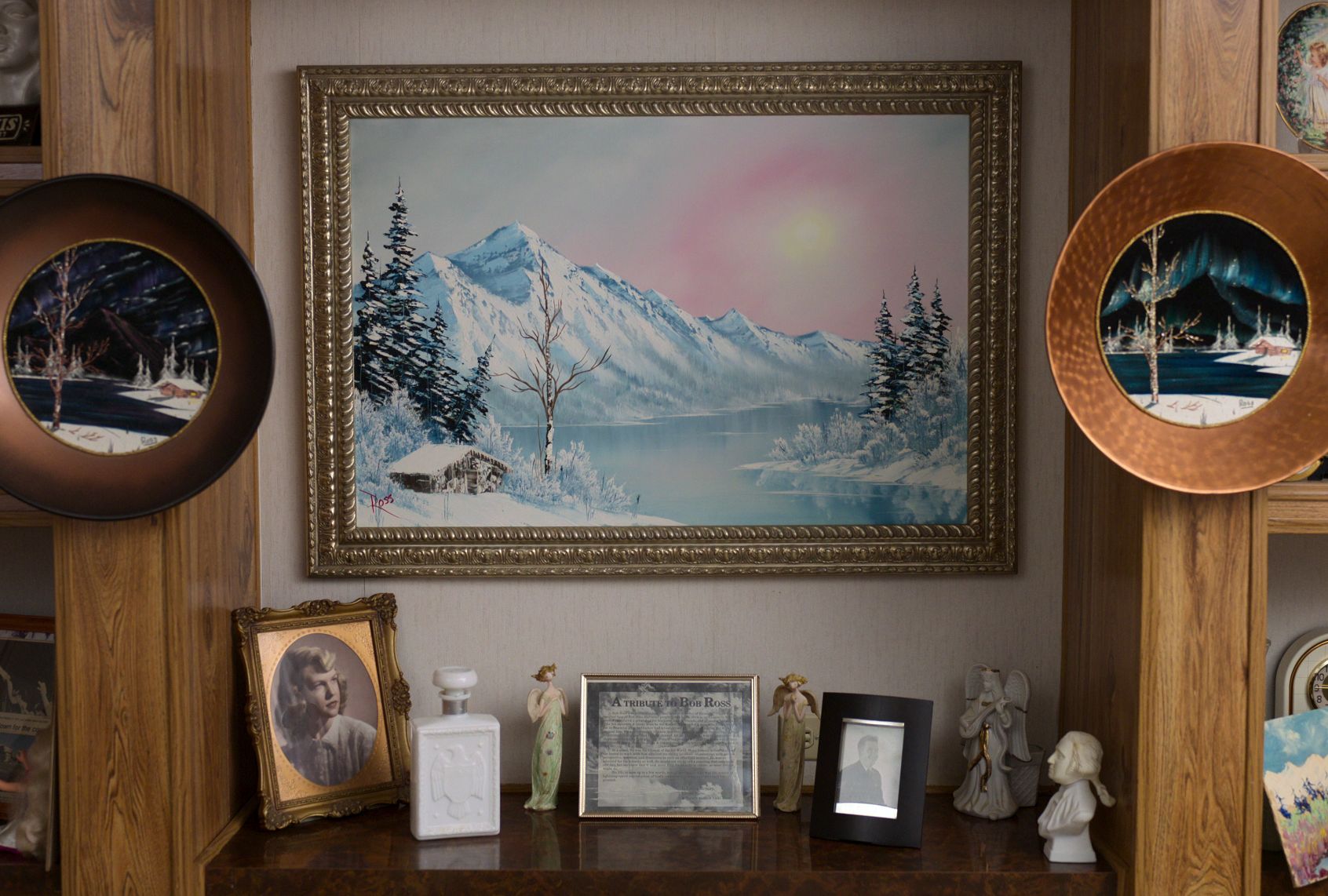 A cosy home alcove displaying various art. A central snowy mountain landscape painting is in the center with two smaller landscapes on plates on either side. Knickknacks and family photos sit on the ledge below