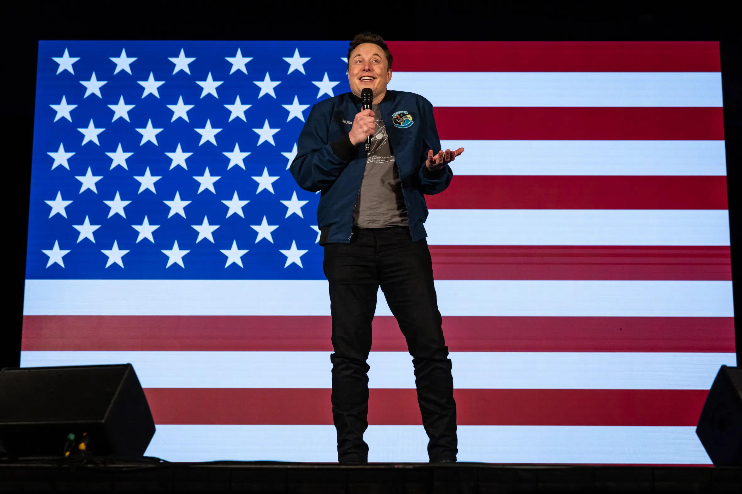 Elon Musk speaks at an America PAC town hall in front of an American flag.
