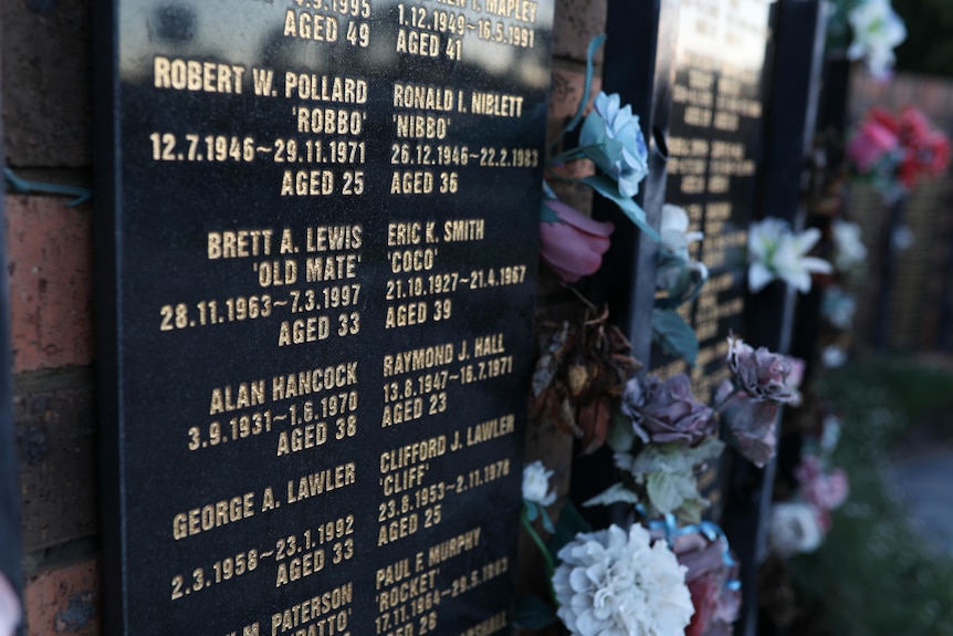 A close up of names, birth and death dates and ages on a memorial, with fake flowers placed around the memorial.