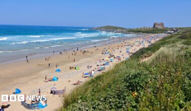 The golden sand beach at Newquay is full of people sunbathing with wind breakers, tents and umbrellas.  The water is busy with swimmers. The Headland Hotel is visible at the far end of the beach.