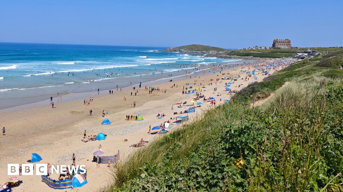 The golden sand beach at Newquay is full of people sunbathing with wind breakers, tents and umbrellas.  The water is busy with swimmers. The Headland Hotel is visible at the far end of the beach.