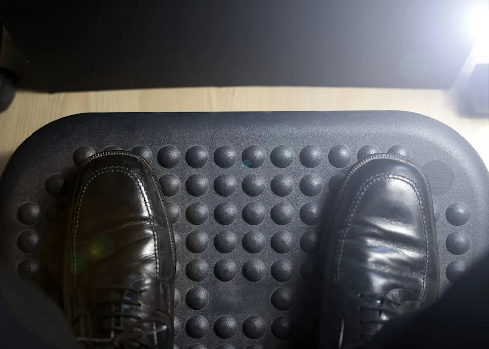 Feet in polished shoes rest on a textured anti-fatigue mat, with round bumps for comfort, on a wooden floor
