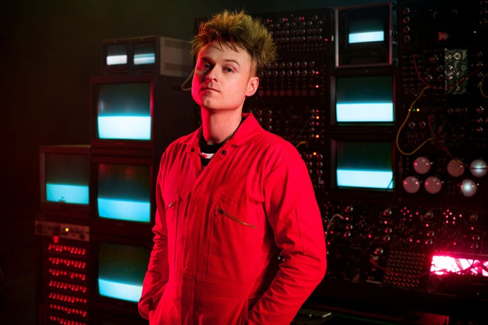 Sam Battle stood next to a coloured background, wearing a red boiler suit.