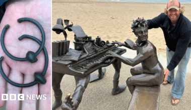 Crowd attends unveiling ceremony on a beach as two people pose for a photograph in front of statue of naked Terry Jones playing a piano